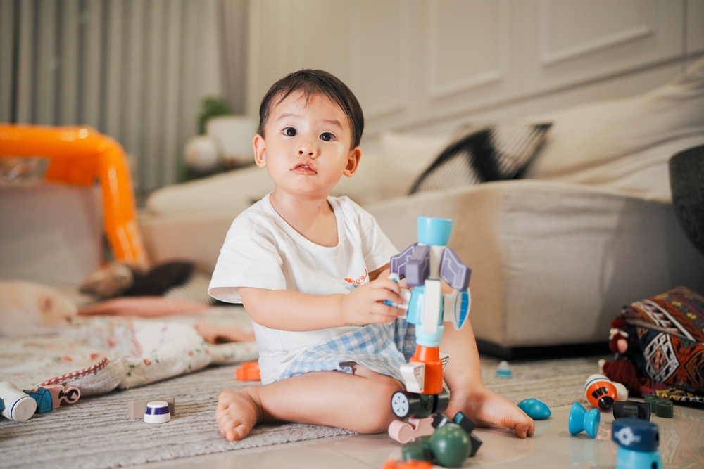 Cute asian toddler sitting on the floor playing with colorful toys in a cozy living room, showcasing joyful childhood moments.