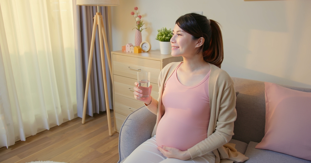 Proper nutrition concept - asian pregnant woman is taking vitamin or medicine with a glass of water in living room at home