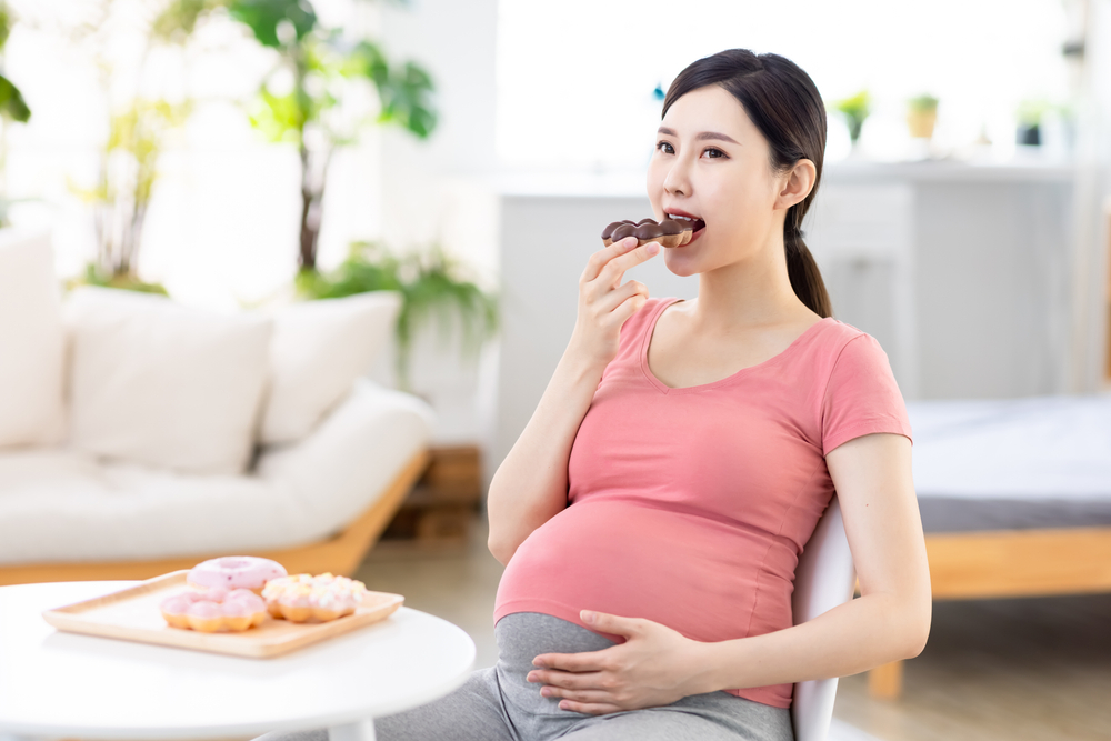 unhealthy eating concept - Asian pregnant woman eats donuts at living room